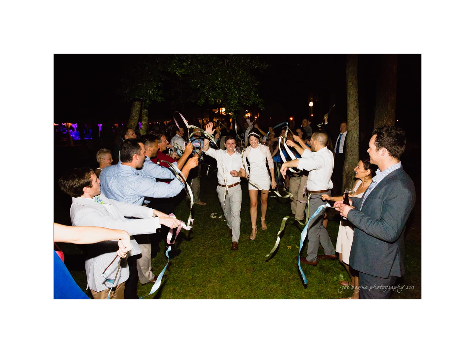 NC wedding photography - rachel & joe A couple smiles and walks hand in hand through an aisle of cheering people waving ribbons. The event appears to be a nighttime celebration, with guests dressed in formal attire and the scene illuminated by ambient lighting.