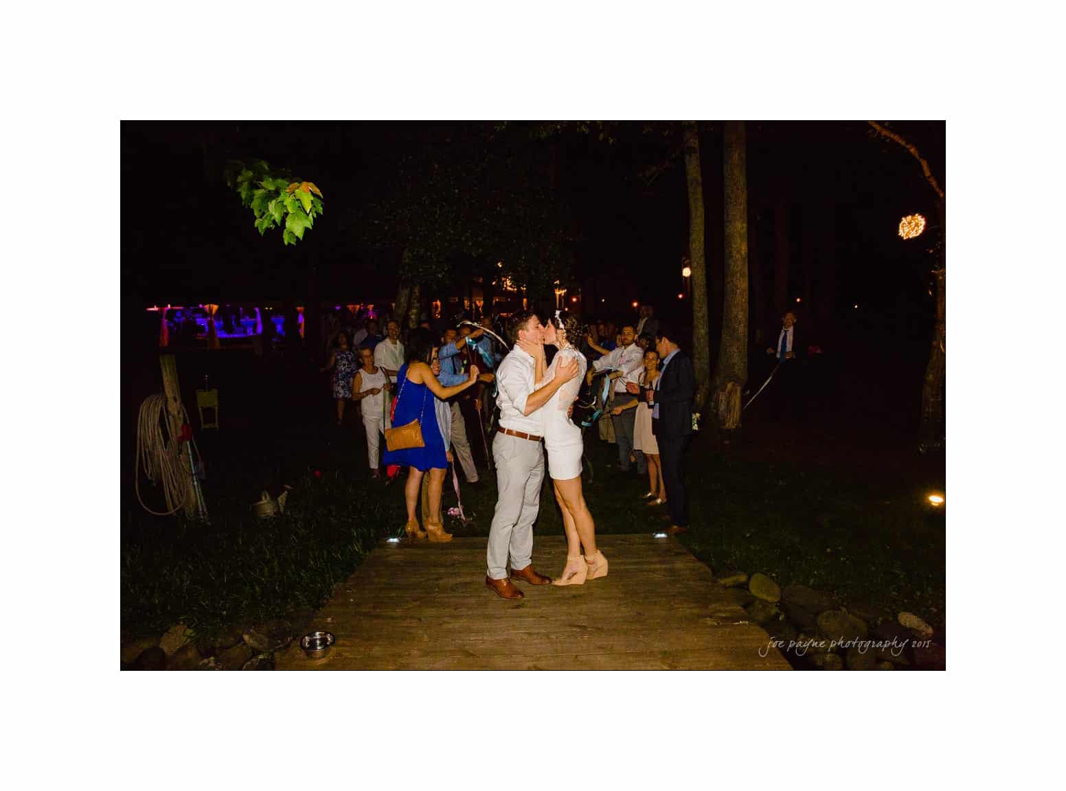 NC wedding photography - rachel & joe A couple is kissing on a wooden path at night, surrounded by a few people. The setting is dark with some people in the background appearing to mingle under subtle lighting. Trees and foliage can be seen around the area.