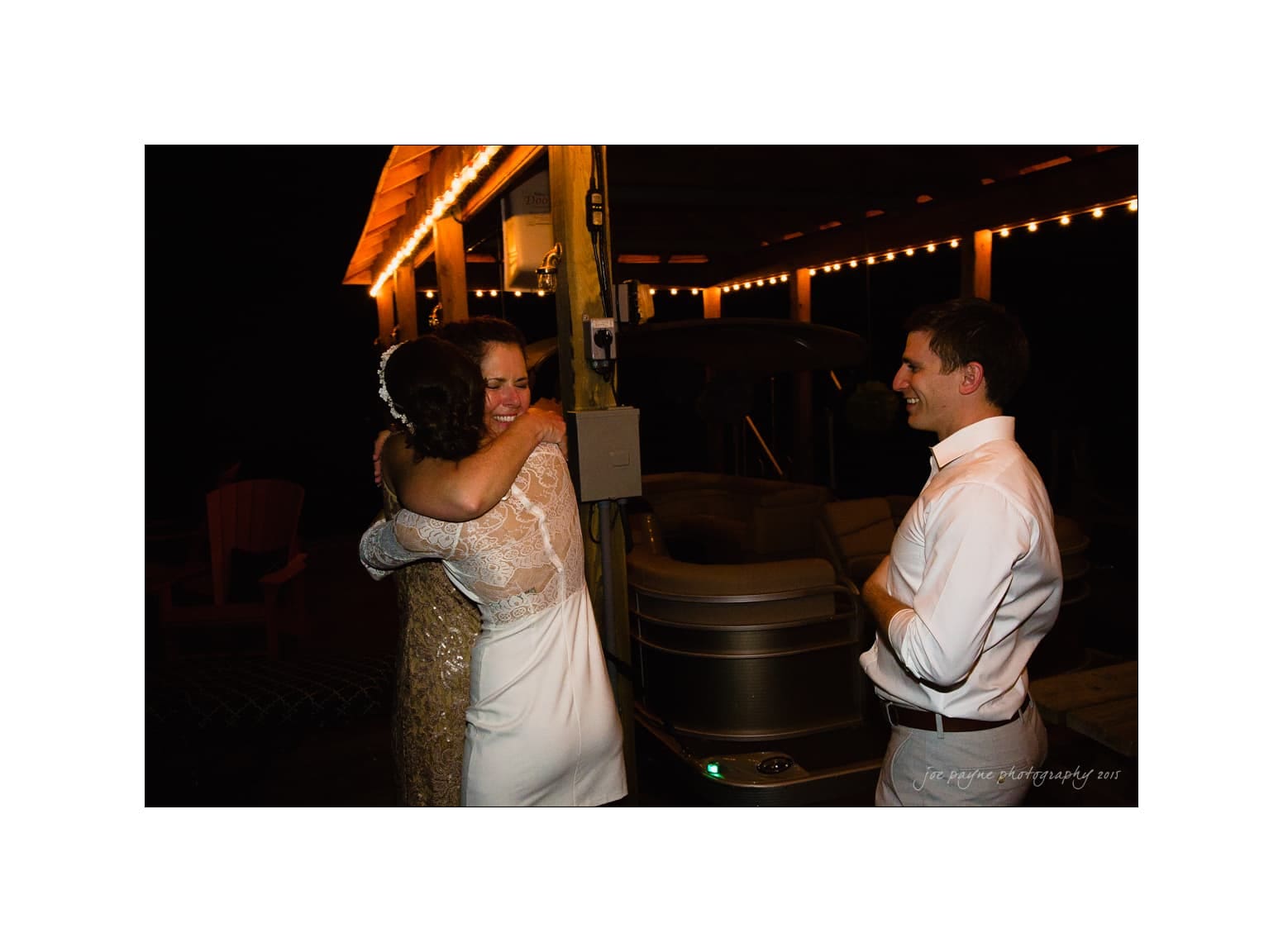 NC wedding photography - rachel & joe A bride in a white dress hugs a woman under a decorated pavilion with string lights at night. A man in a white shirt stands nearby, smiling at the scene.