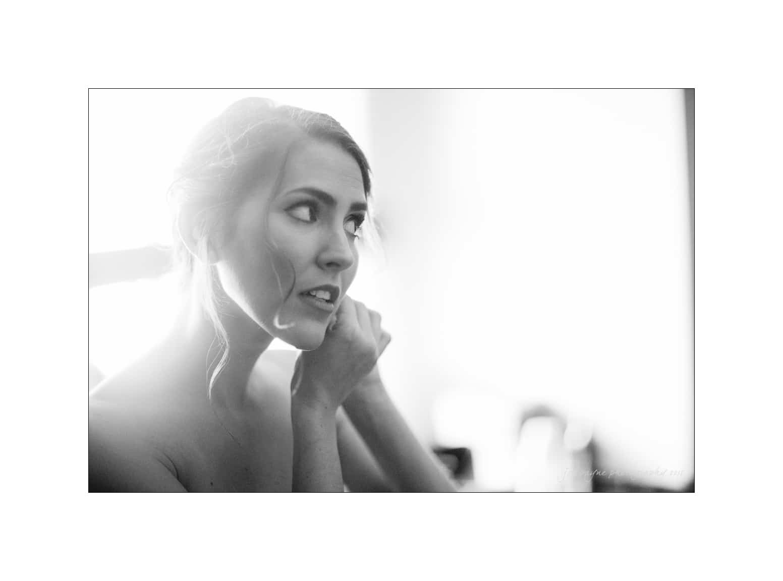NC wedding photography - rachel & joe A woman in a strapless dress adjusts her earring while looking to the side. The photo is in black and white, with soft lighting and a blurred background, highlighting her thoughtful expression.