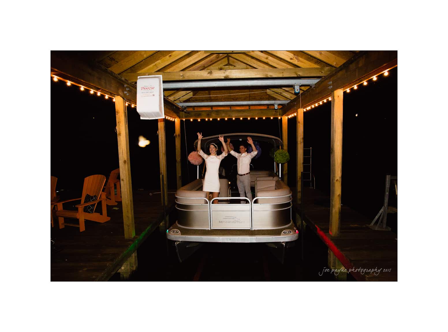 NC wedding photography - rachel & joe A couple stands on a pontoon boat under a wooden canopy decorated with string lights. Both have their arms raised in celebration. The dock is lined with Adirondack chairs. The scene is festive and nighttime.