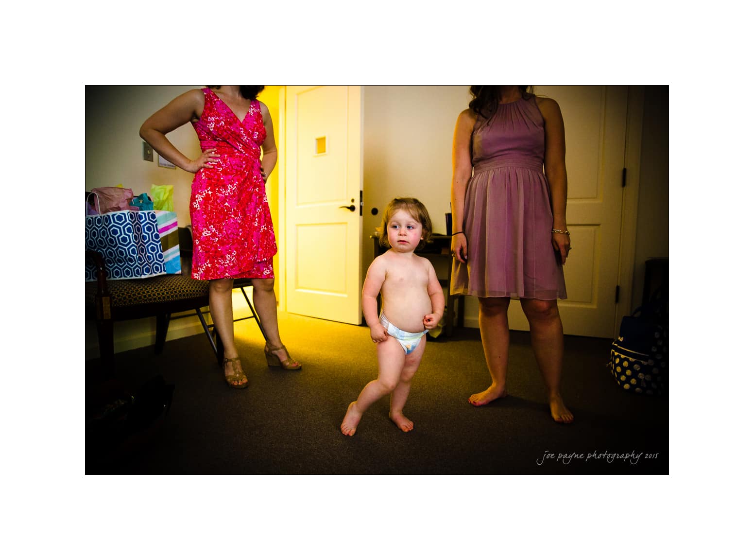 NC wedding photography - rachel & joe A toddler in a diaper stands in a room with two women, one in a pink dress and the other in a purple dress. Shopping bags are visible on a chair, and the room is dimly lit with cream-colored walls.