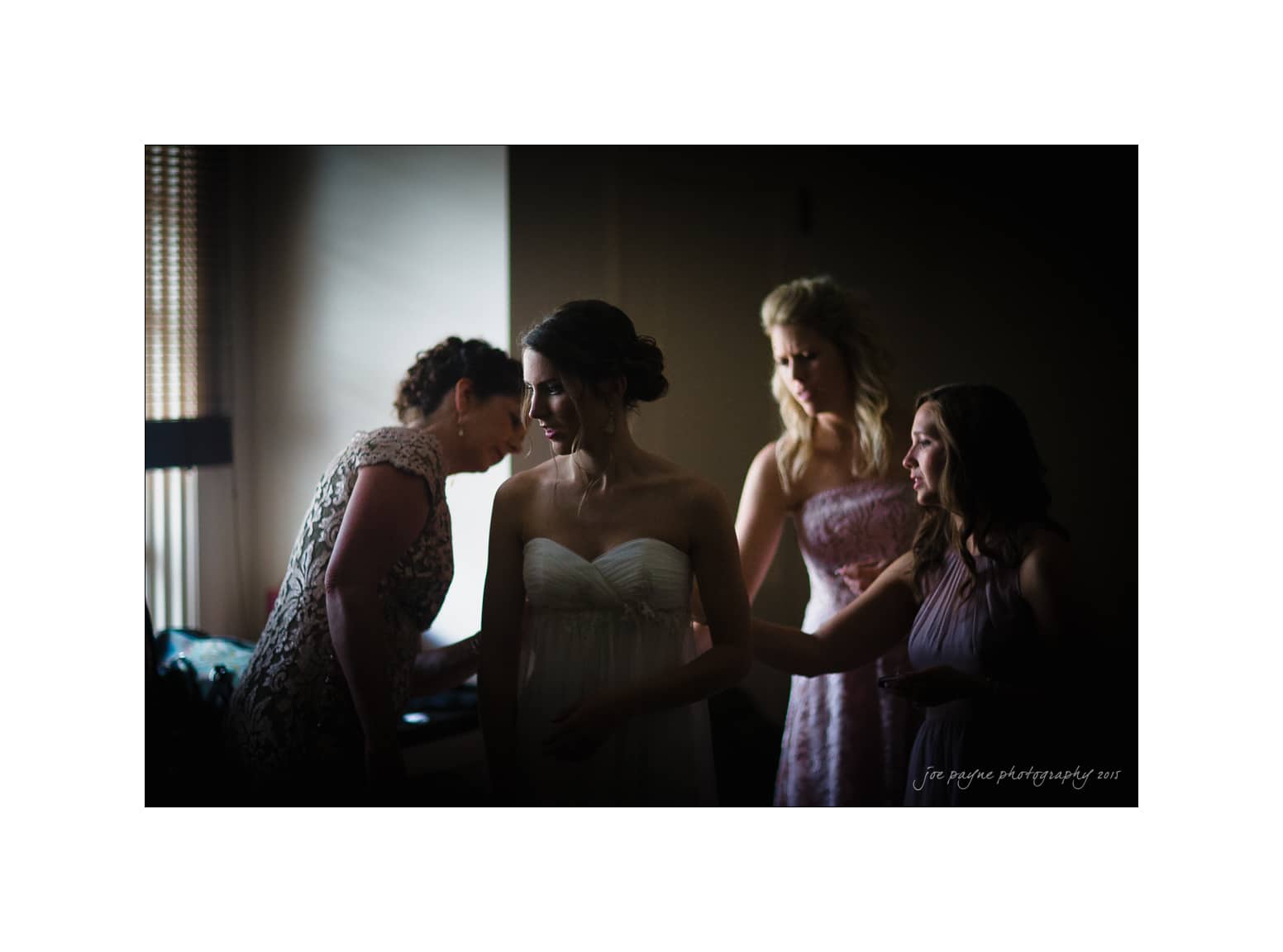 NC wedding photography - rachel & joe A group of four women in a dimly lit room, with one woman in a white strapless dress standing in the center. The others, wearing bridesmaid dresses, are attending to her. The atmosphere is calm and focused.