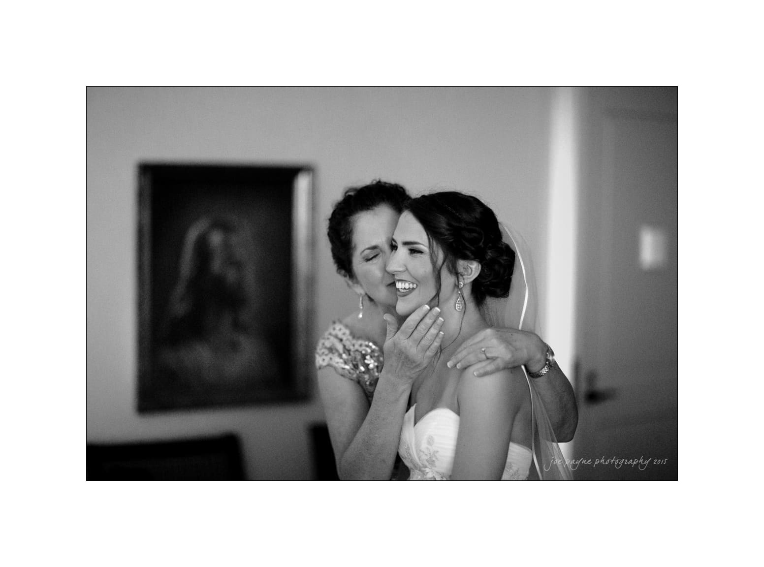 NC wedding photography - rachel & joe A black and white photo of a joyful bride being embraced by an older woman indoors. They are both smiling warmly. A portrait of a man hangs on the wall in the background.
