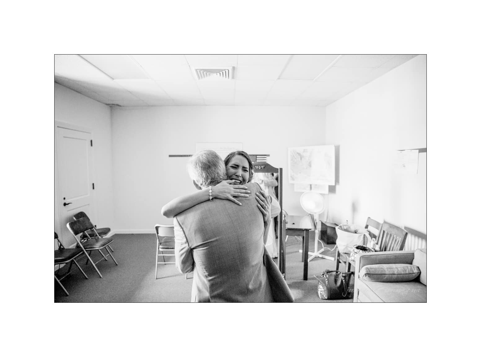 NC wedding photography - rachel & joe A black and white photo of two people embracing in a small room. One person is facing the camera with a smile, while the other has their back to the camera. Chairs and a map are in the background.
