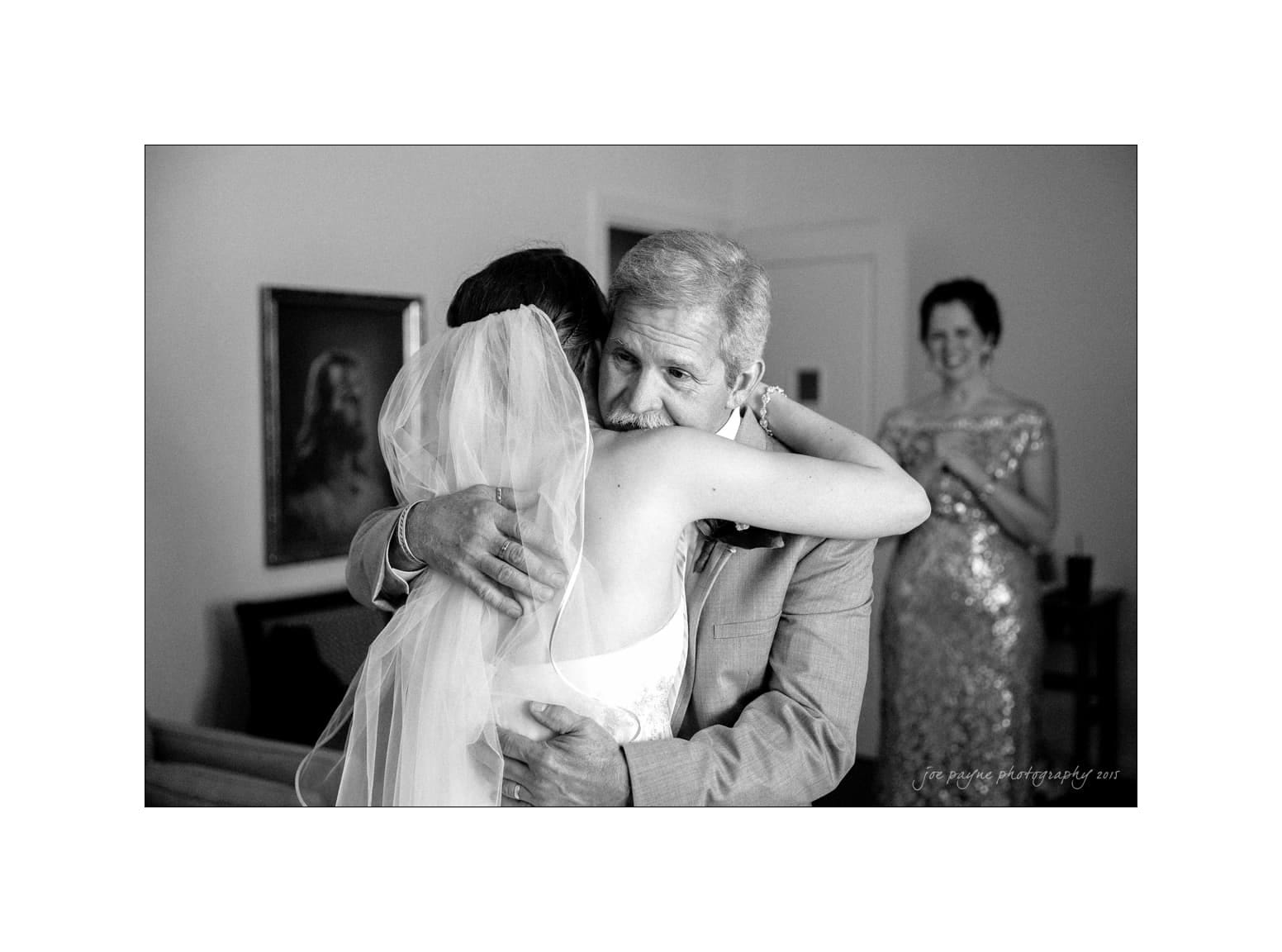 NC wedding photography - rachel & joe A bride in a veil shares an emotional hug with an older man in a suit. A smiling woman in an elegant dress watches in the background. The room is softly lit, and a portrait hangs on the wall.
