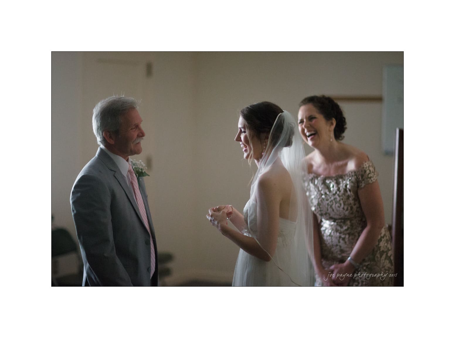 NC wedding photography - rachel & joe A bride in a wedding dress and veil laughs with two well-dressed individuals in a softly lit room. One person wears a light-colored suit, and the other dons an elegant patterned dress. The atmosphere is joyful and celebratory.