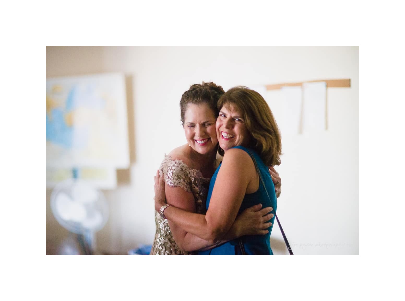 NC wedding photography - rachel & joe Two women hugging and smiling warmly. The woman on the left is wearing a sparkly dress, and the woman on the right is in a blue dress. They appear to be in a warmly lit room with soft-focus artwork in the background.