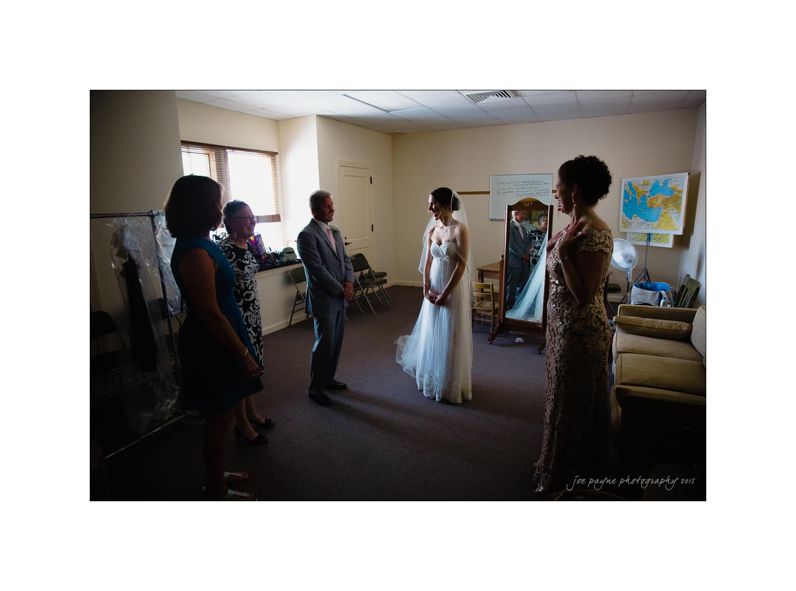 NC wedding photography - rachel & joe A bride stands in a room with four people, wearing a white wedding dress. Two women and two men are around her. The room is softly lit, with a map and a sofa in the background. A dress bag hangs to the side.