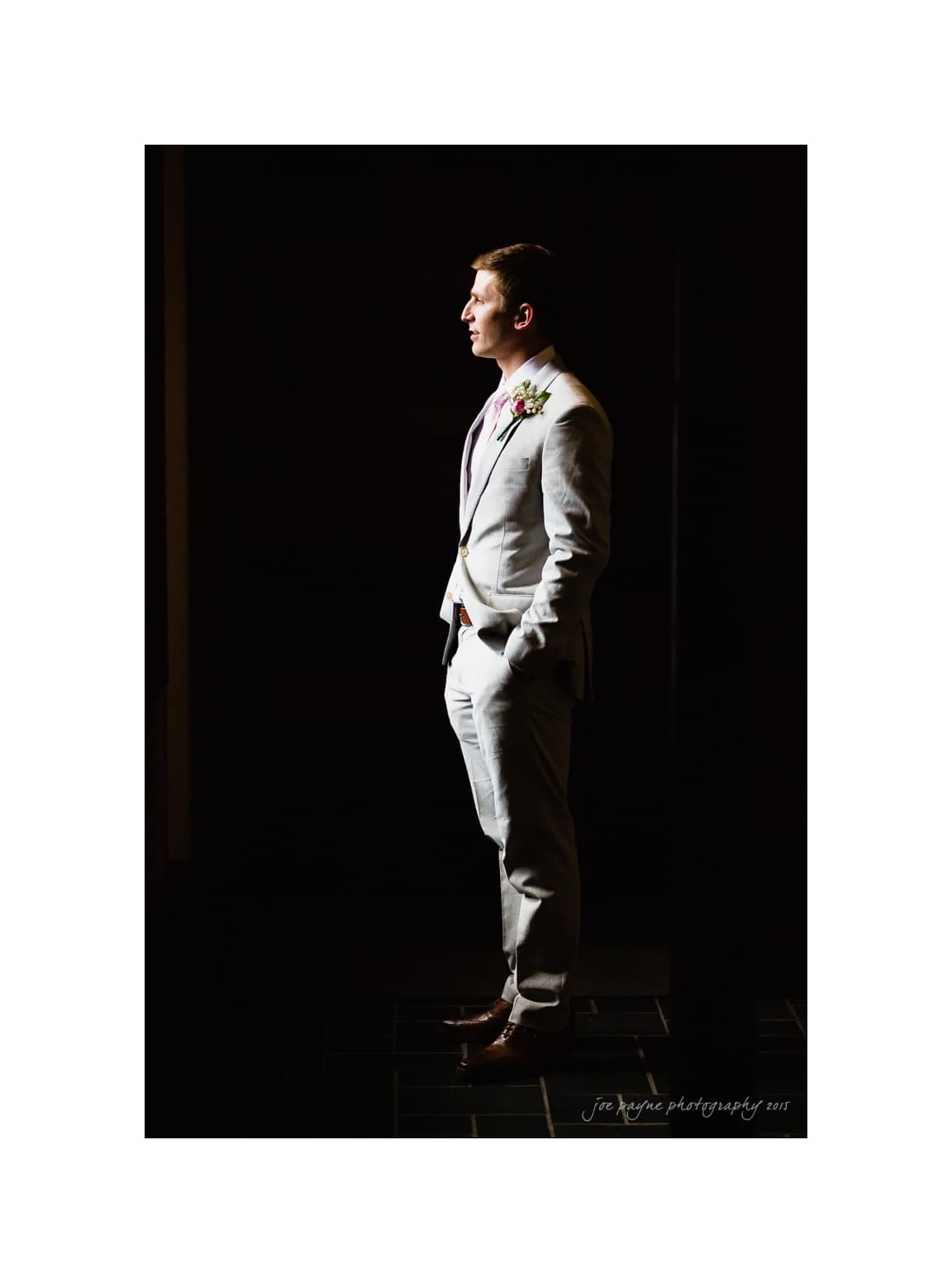NC wedding photography - rachel & joe A man in a light gray suit stands in profile, hands in pockets, looking thoughtfully into the light. Hes wearing a boutonniere, and the background is dark, emphasizing the spotlight on him.