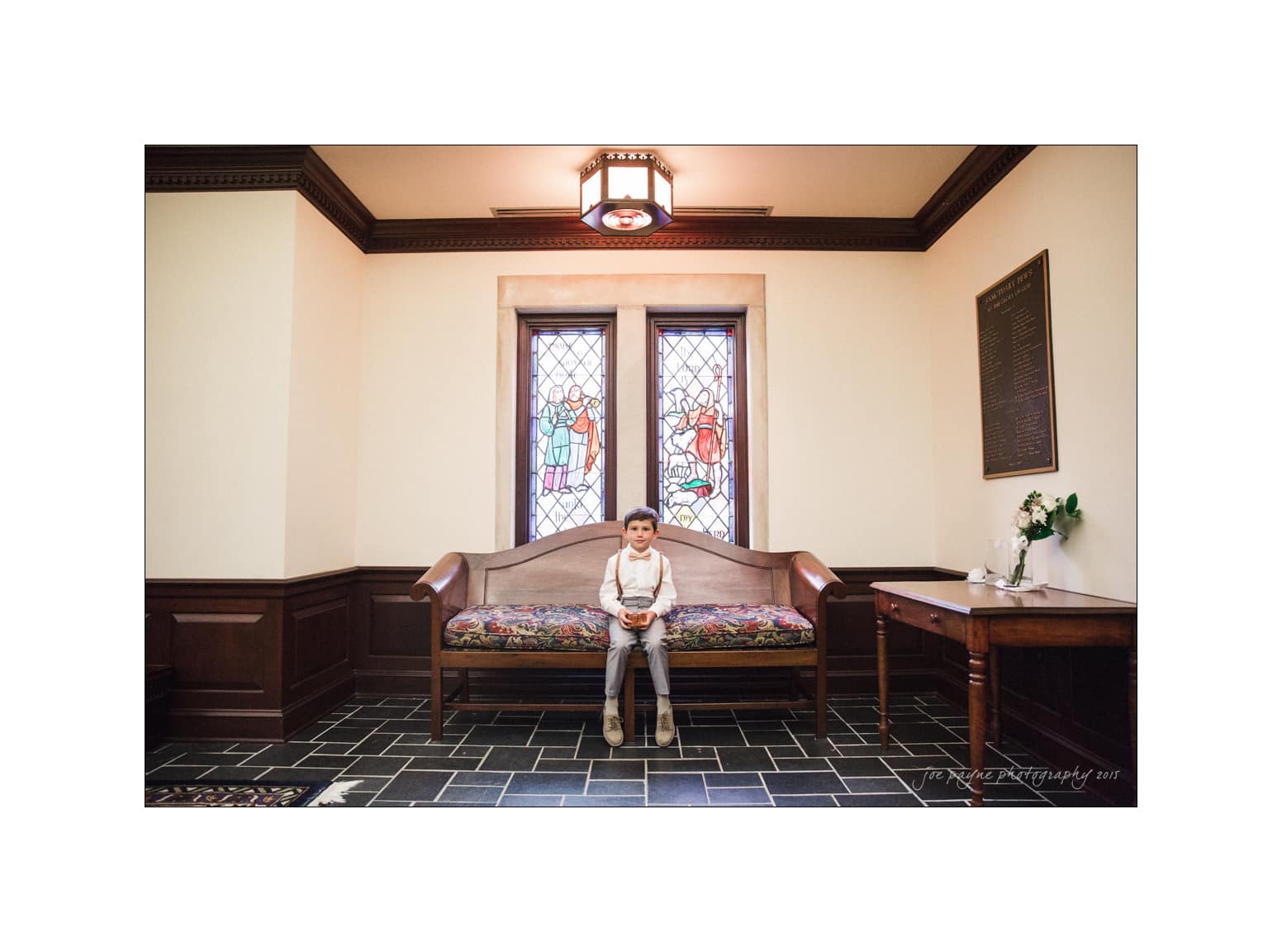 NC wedding photography - rachel & joe A child sits on a wooden bench in a church, facing the camera. Behind the child are two colorful stained glass windows depicting figures. The room has beige walls, dark wood paneling, and a small side table with a floral arrangement.