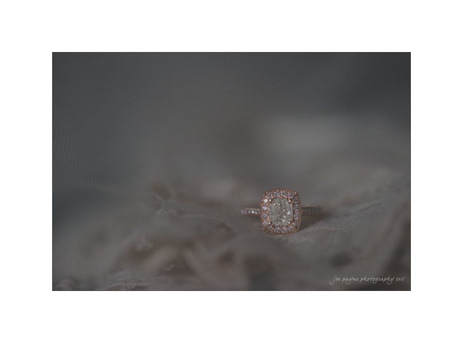 NC wedding photography - rachel & joe Close-up of a diamond ring with a square-shaped setting and small stones surrounding the center diamond. The ring is placed on a soft, textured fabric background.