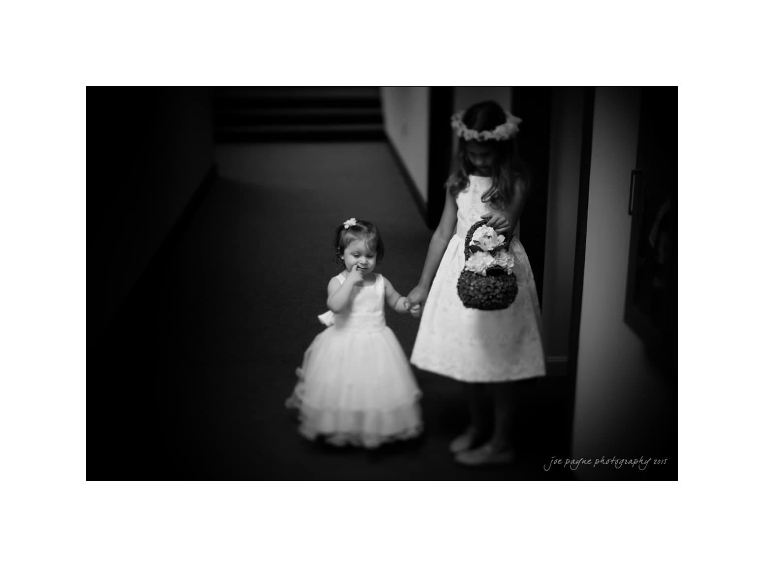 NC wedding photography - rachel & joe A black and white photo of two young girls in dresses. The older girl wears a flower crown and holds a basket, while holding hands with the younger girl, who is in a similar dress. They stand in a dimly lit hallway.
