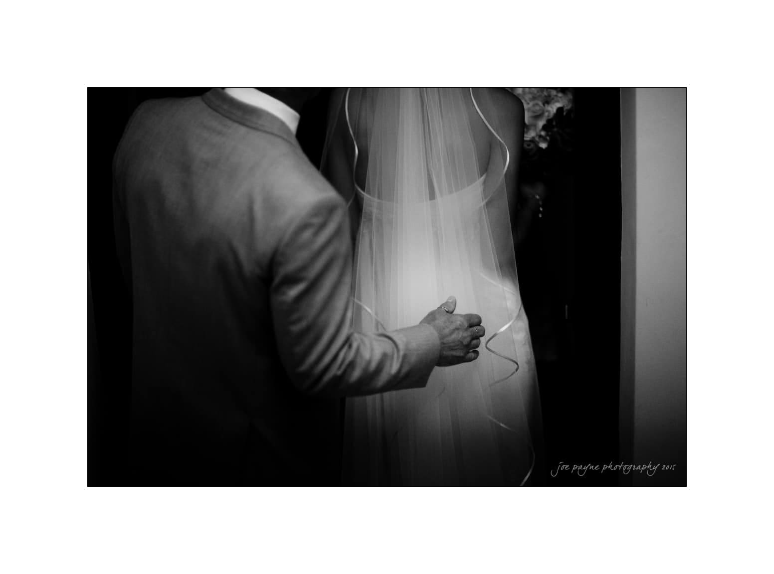 NC wedding photography - rachel & joe A black-and-white image of a groom gently placing his hand on the back of a bride, whose back is turned. The bride is wearing a flowing veil and dress. The scene captures an intimate moment during their wedding.