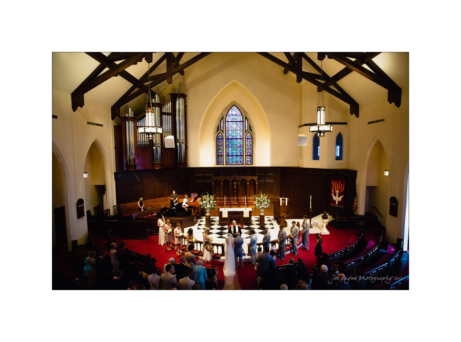 NC wedding photography - rachel & joe A wedding ceremony in a church with a high ceiling and stained glass window, featuring a bride and groom at the altar surrounded by the bridal party and guests seated in pews. The organ and floral arrangements decorate the space.