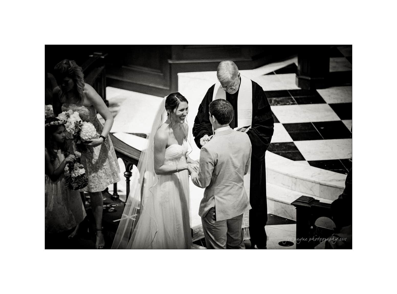 NC wedding photography - rachel & joe A black and white photo of a wedding ceremony. A couple stands facing each other, the bride in a white gown and the groom in a suit. An officiant in a robe stands behind them. Bridesmaids with bouquets stand to the side.