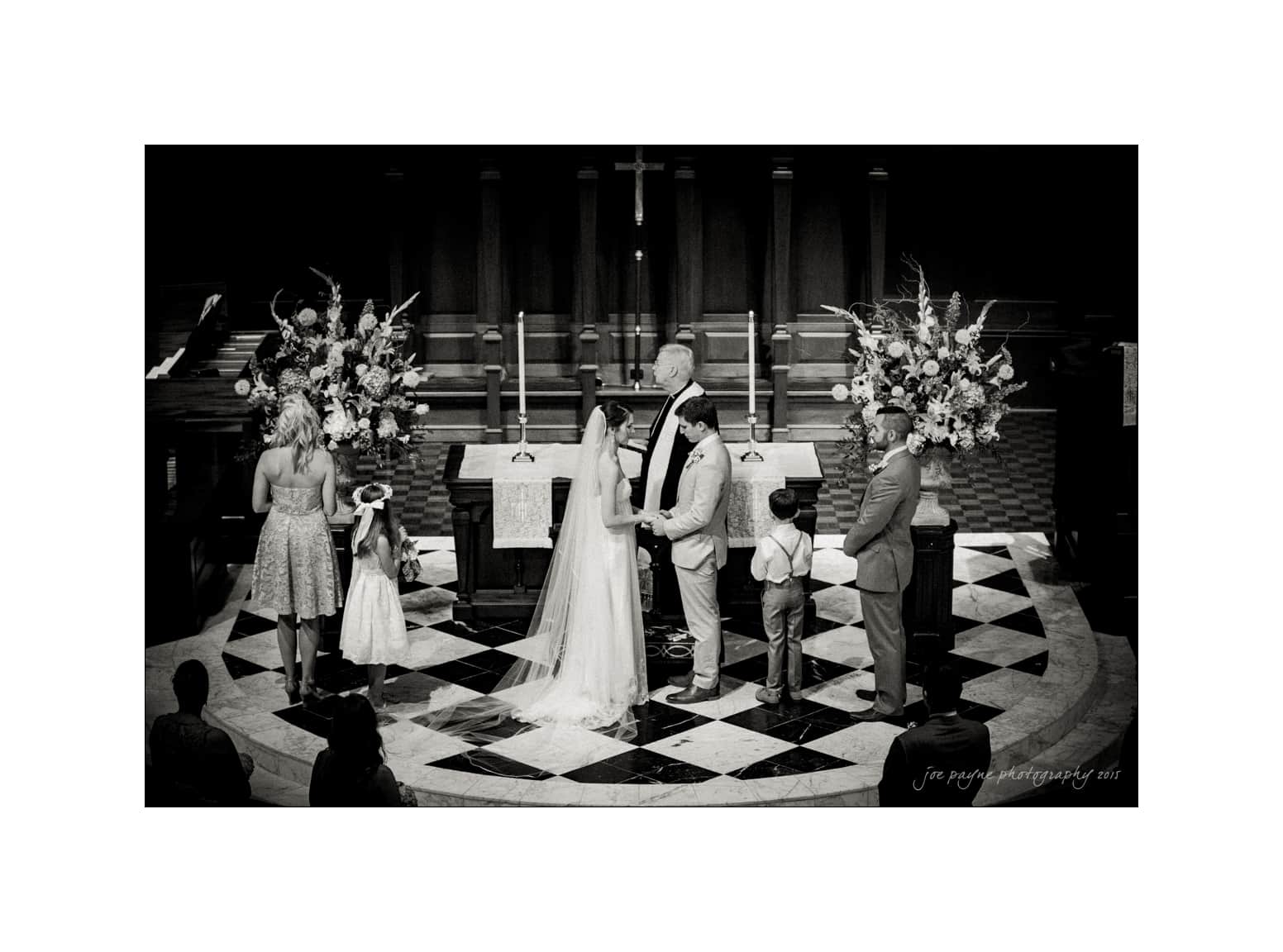 NC wedding photography - rachel & joe A black and white image of a wedding ceremony. The bride and groom stand facing each other at the altar, surrounded by the bridal party and two children. Large floral arrangements are on either side of the couple.