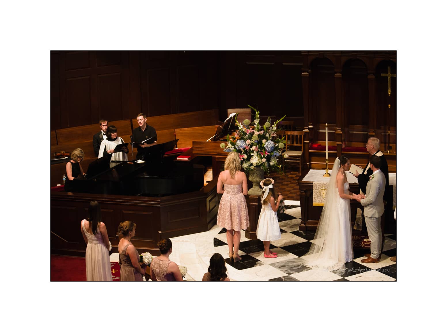 NC wedding photography - rachel & joe A wedding ceremony in a church shows a couple holding hands at the altar. A young flower girl stands nearby, and a pianist and singers perform. Bridesmaids in light dresses stand to one side, with a large floral arrangement in the background.