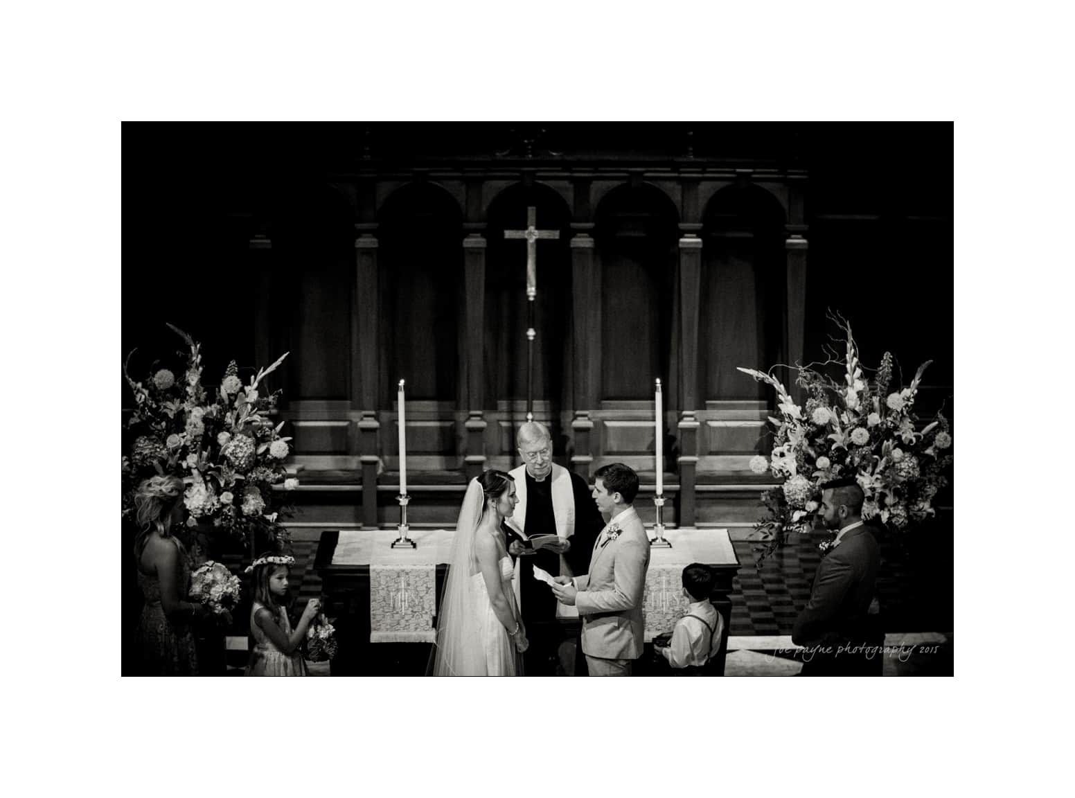 NC wedding photography - rachel & joe A bride and groom stand at the altar, holding hands during their wedding ceremony in a church. The officiant stands behind them, and bridesmaids and groomsmen are visible. Large floral arrangements decorate the altar.