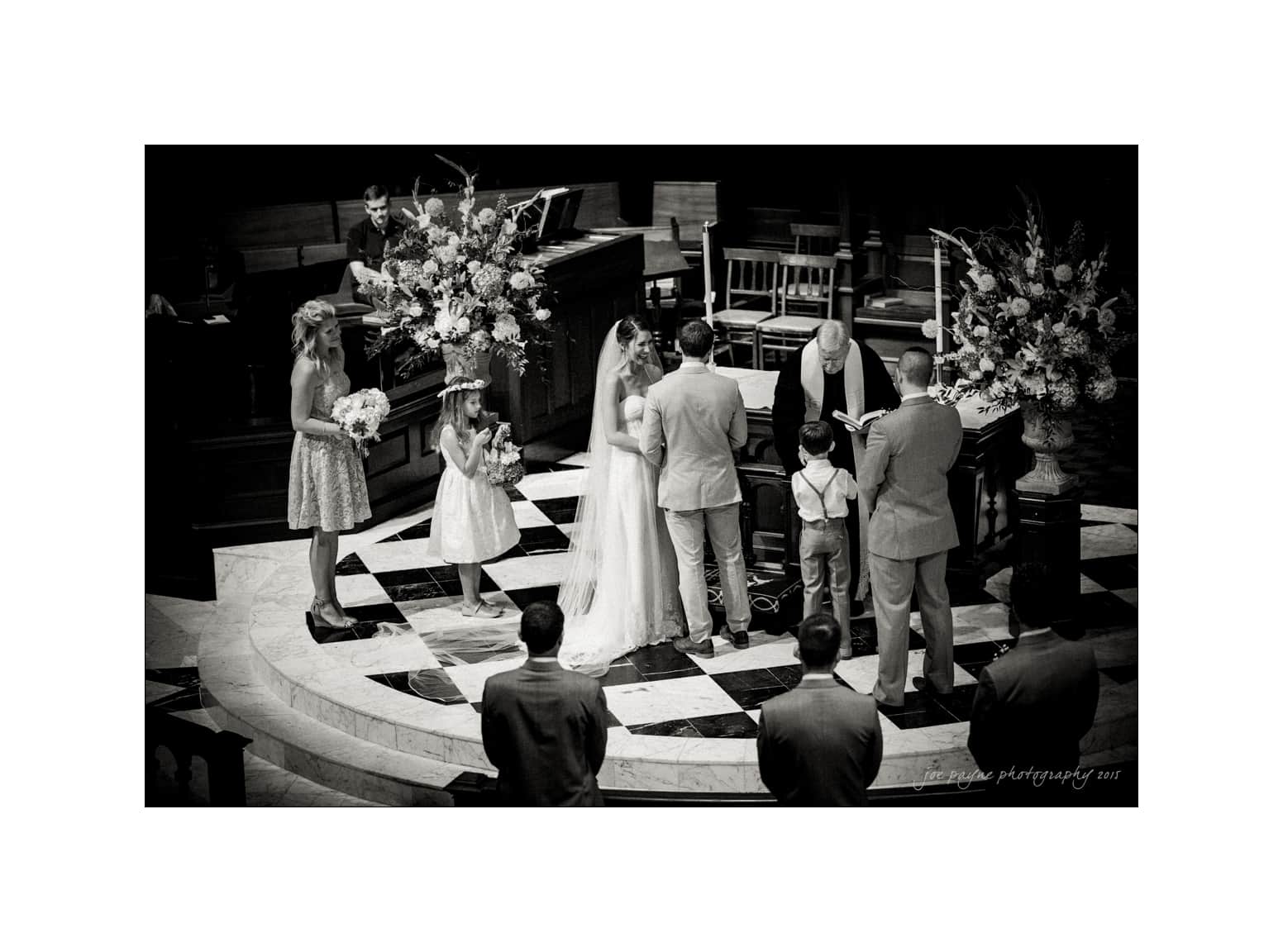 NC wedding photography - rachel & joe A black and white photo of a wedding ceremony in a church. The bride and groom stand facing each other with the officiant in front of them. A flower girl and ring bearer are nearby, with guests observing. Large floral arrangements decorate the space.