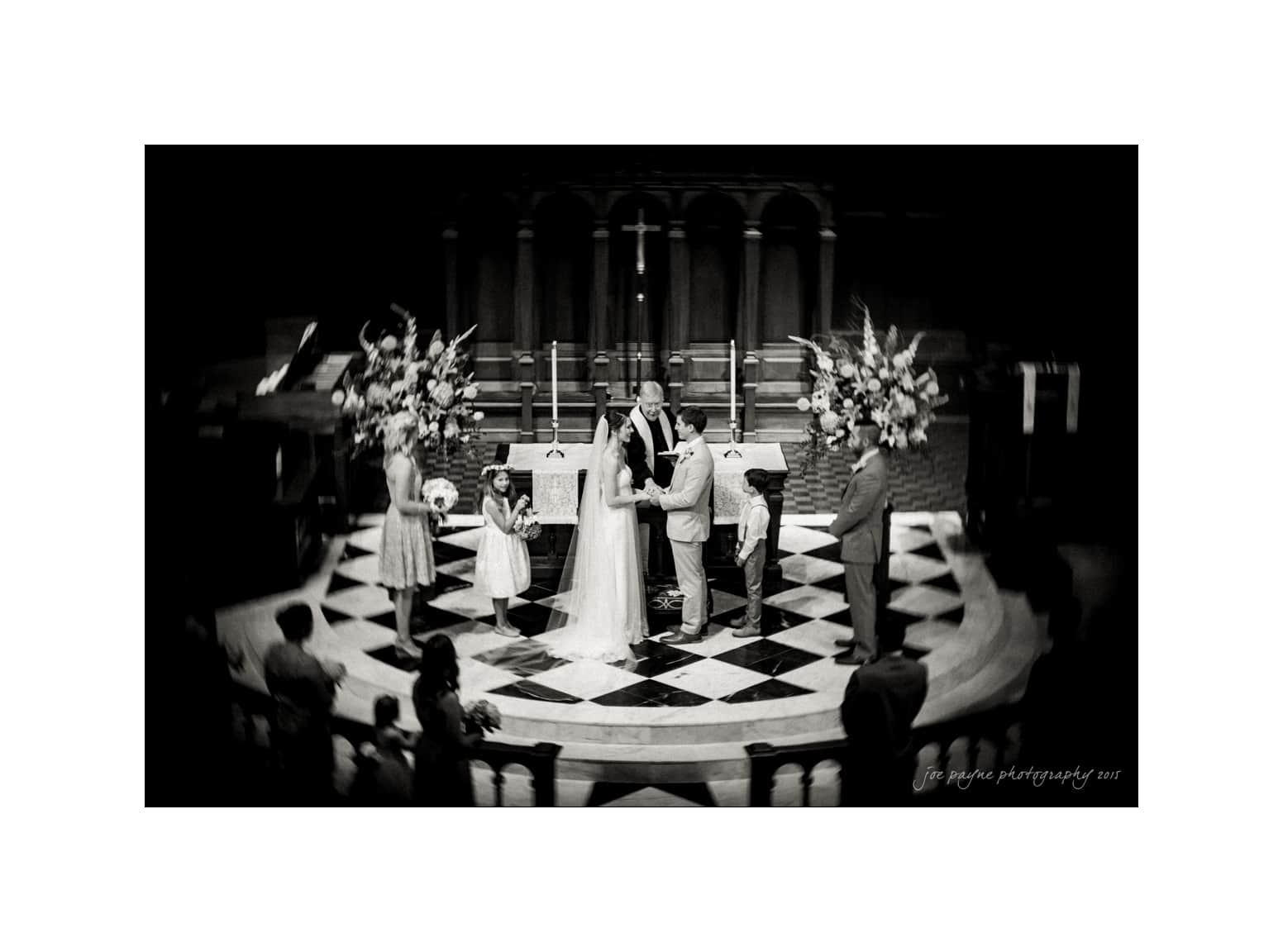 NC wedding photography - rachel & joe A black and white photo of a wedding ceremony. The bride and groom stand at the altar, exchanging vows. Two children stand nearby, possibly as attendants. Large floral arrangements are visible, and the setting appears to be a church.