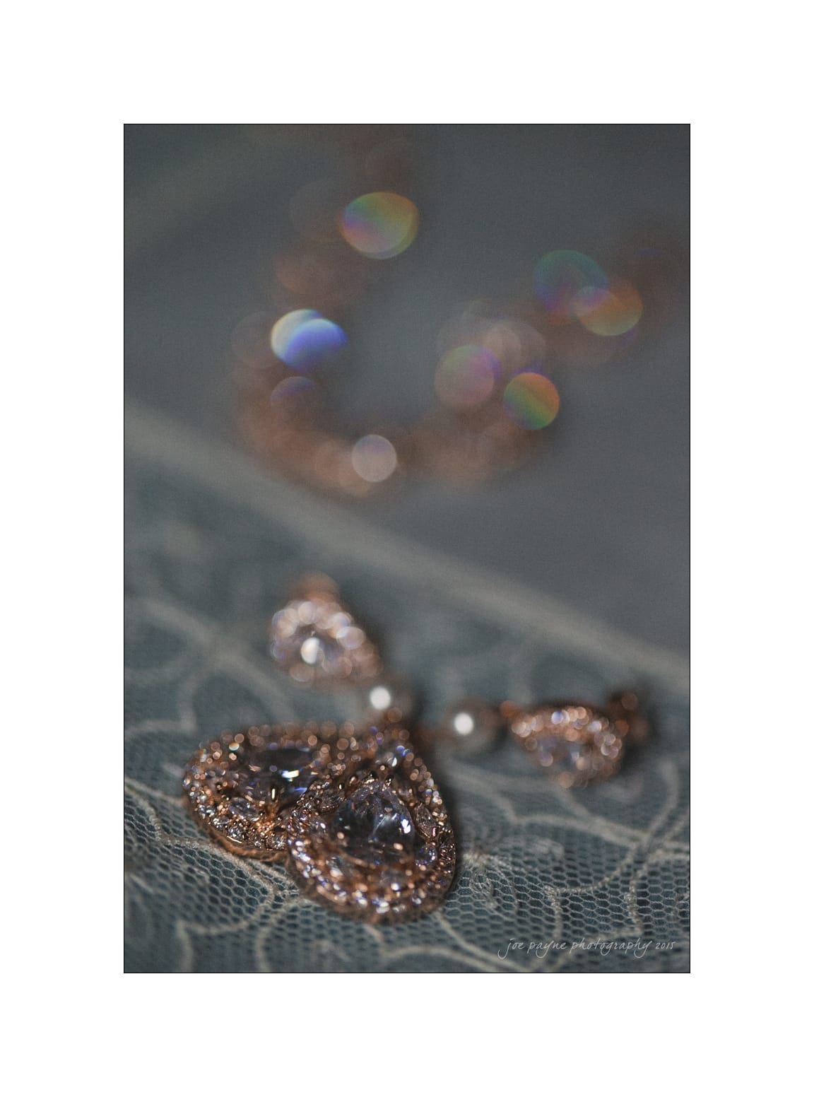 NC wedding photography - rachel & joe Close-up of a pair of ornate teardrop earrings with sparkling stones, resting on a textured lace fabric. A blurred necklace is visible in the background, adding a touch of colorful bokeh.