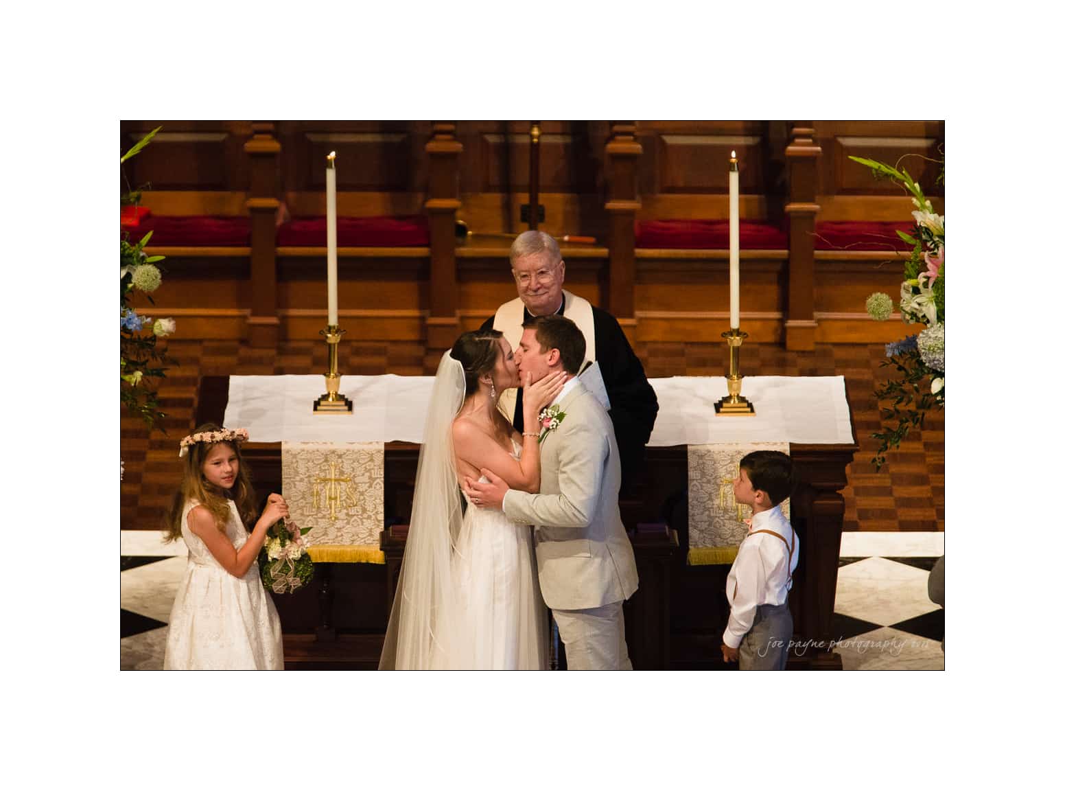 NC wedding photography - rachel & joe A bride and groom share a kiss at the altar during a wedding ceremony. The officiant stands behind them, and two children, a flower girl and a ring bearer, are nearby. Candles and floral decorations adorn the setting.