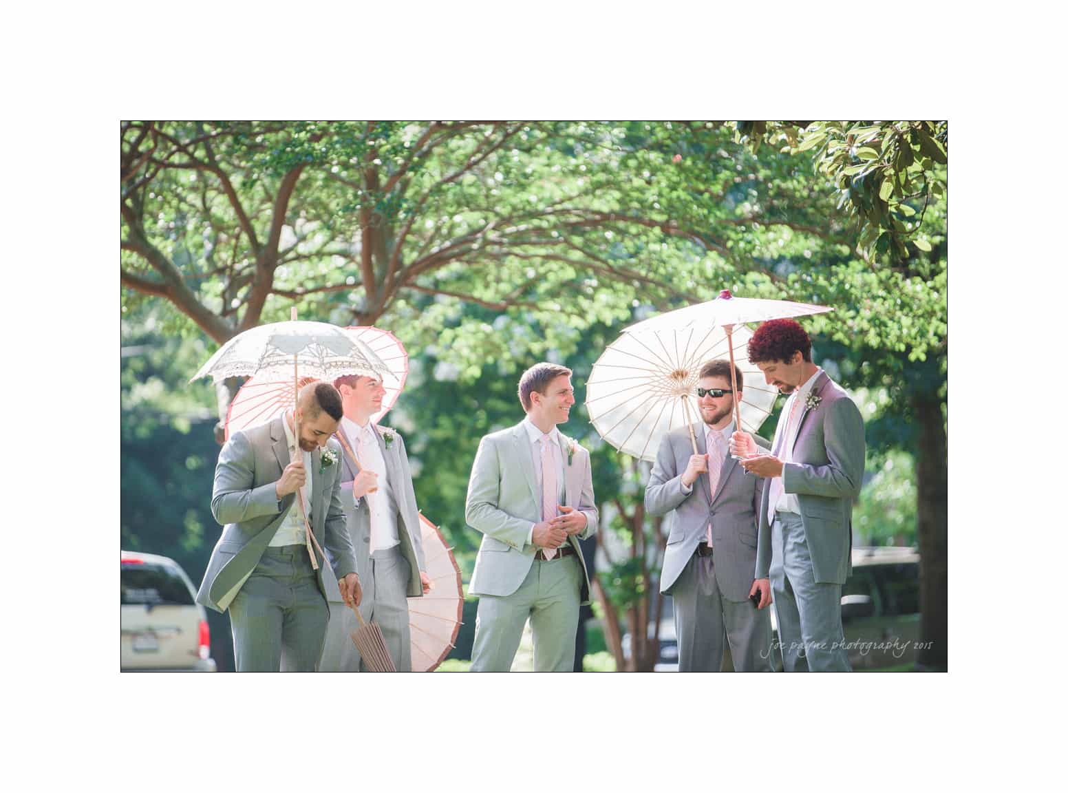 NC wedding photography - rachel & joe A group of five men in light gray suits stand outdoors under dappled sunlight. Some hold lace and paper parasols. Trees and greenery form the background, and a vehicle is partially visible on the left.
