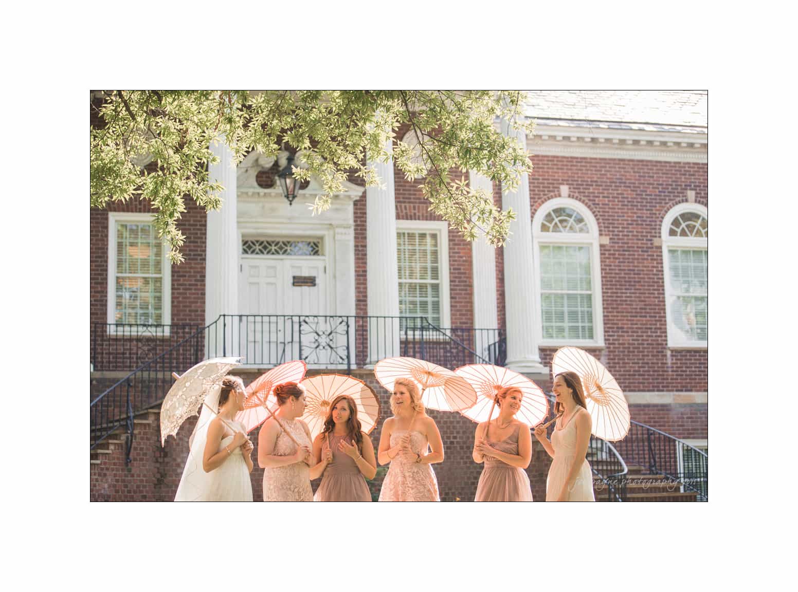 NC wedding photography - rachel & joe A group of six women in dresses stand in front of a brick building with white columns, holding decorative umbrellas. They are on a sidewalk surrounded by greenery. The scene is bright and sunny.