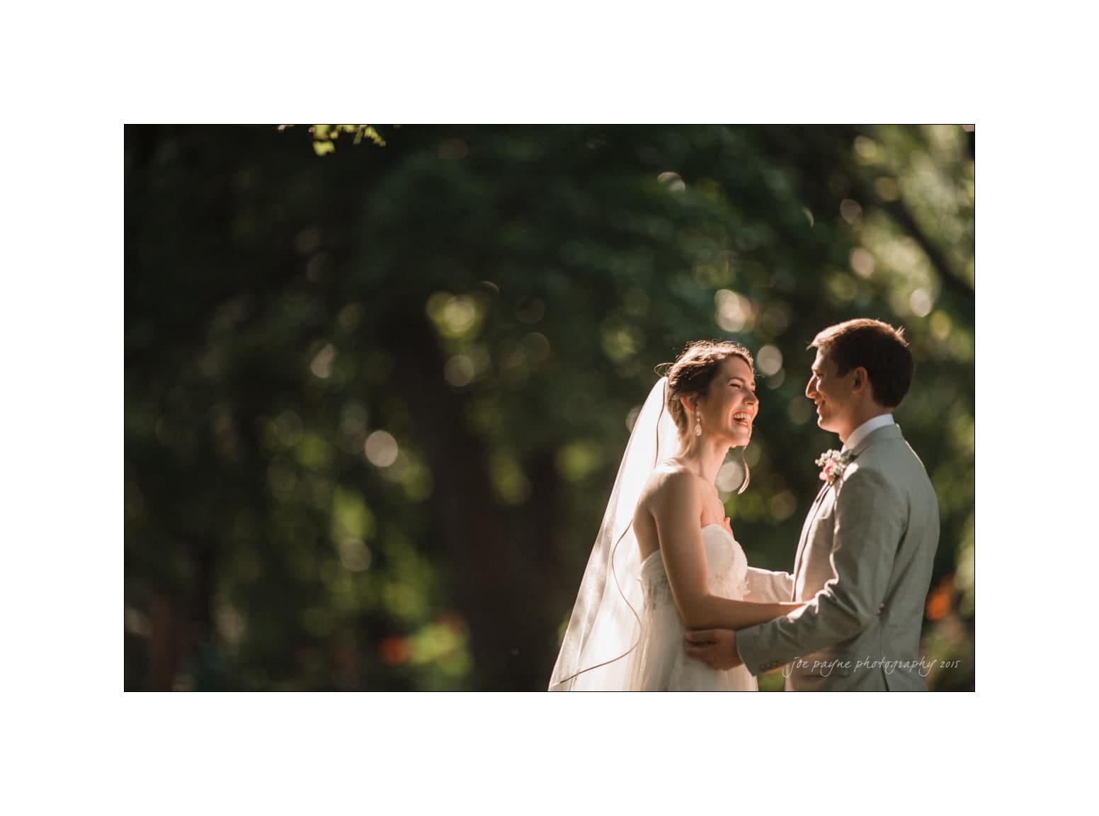 NC wedding photography - rachel & joe A bride and groom stand facing each other, smiling, in a sunlit forest setting. The bride wears a veil, and the groom is in a light-colored suit. Dappled sunlight filters through the trees, creating a romantic atmosphere.
