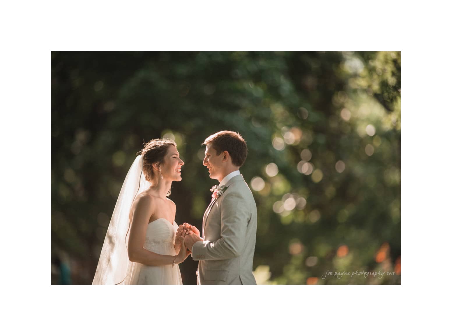 A bride and groom stand facing each other, holding hands and smiling, surrounded by a softly blurred natural background with dappled sunlight. The bride wears a white dress and veil, and the groom is in a light-colored suit.