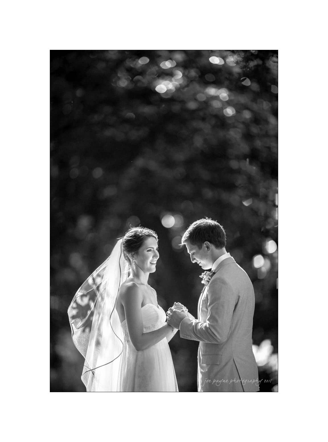 NC wedding photography - rachel & joe A bride and groom stand facing each other, holding hands, in a black and white photograph. The bride is in a wedding dress with a veil, while the groom wears a suit. They are outdoors with soft, blurred lights in the background.