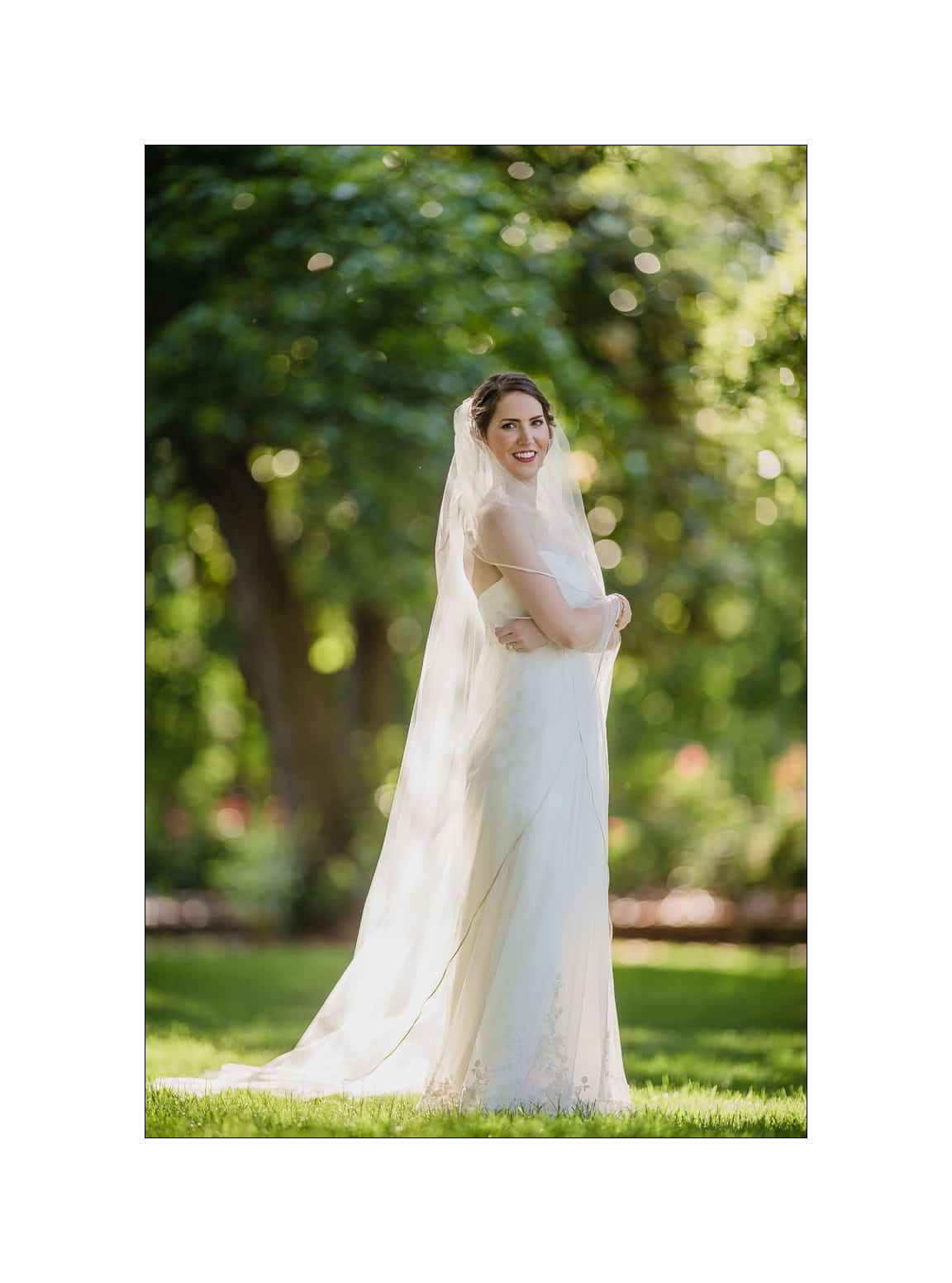 NC wedding photography - rachel & joe A bride in a white dress and veil stands smiling in a sunlit garden. The lush green foliage and soft bokeh create a serene and romantic atmosphere.