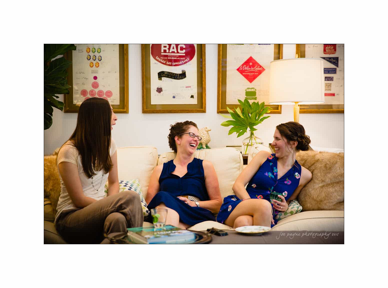 NC wedding photography - rachel & joe Three women sit on a couch, laughing and talking in a cozy, well-decorated room. They are surrounded by framed posters and plants, creating a relaxed and cheerful atmosphere.