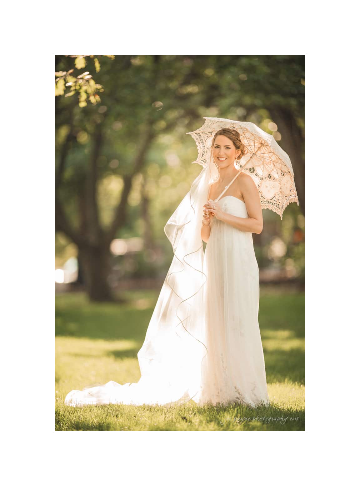 NC wedding photography - rachel & joe A bride in a white dress stands smiling under a lace parasol. Her veil flows down, and she is surrounded by green trees and sunlight in a park setting.