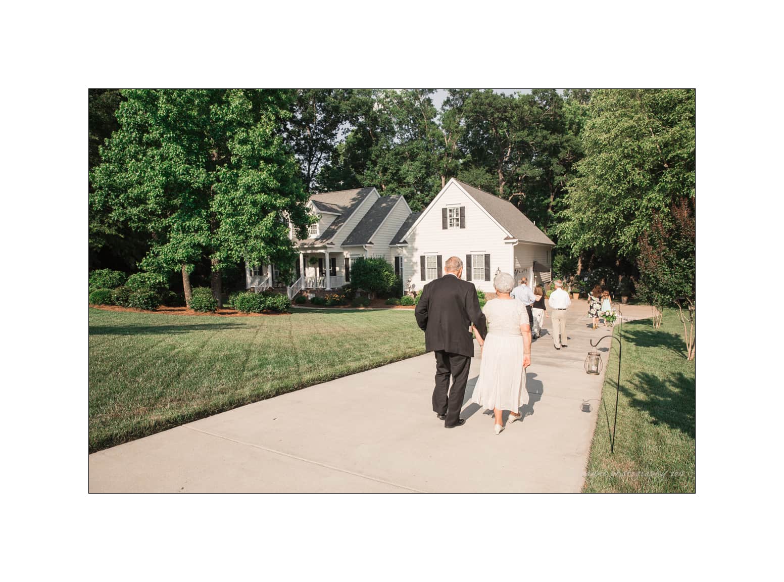 NC wedding photography - rachel & joe An elderly couple walks arm in arm down a driveway toward a charming white house surrounded by lush greenery. They are followed by a few other people, enjoying a sunny day.