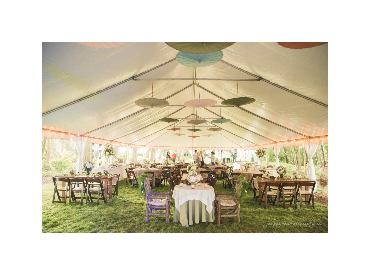 NC wedding photography - rachel & joe A large outdoor tent is set up for an event, with round tables covered in white cloths and adorned with centerpieces. Wooden chairs surround the tables. Colorful paper umbrellas hang from the tents ceiling, and soft lighting creates a warm atmosphere.