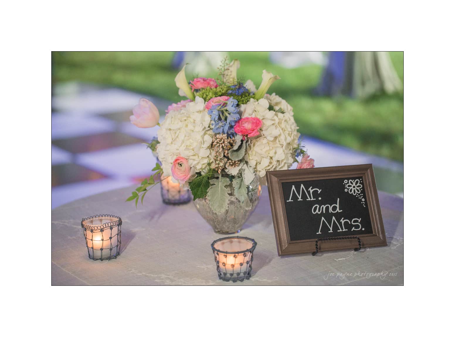 NC wedding photography - rachel & joe A decorative wedding table with a bouquet of flowers, including hydrangeas and tulips, and a small framed chalkboard sign reading Mr. and Mrs. Three lit candles in glass holders are arranged around the display.