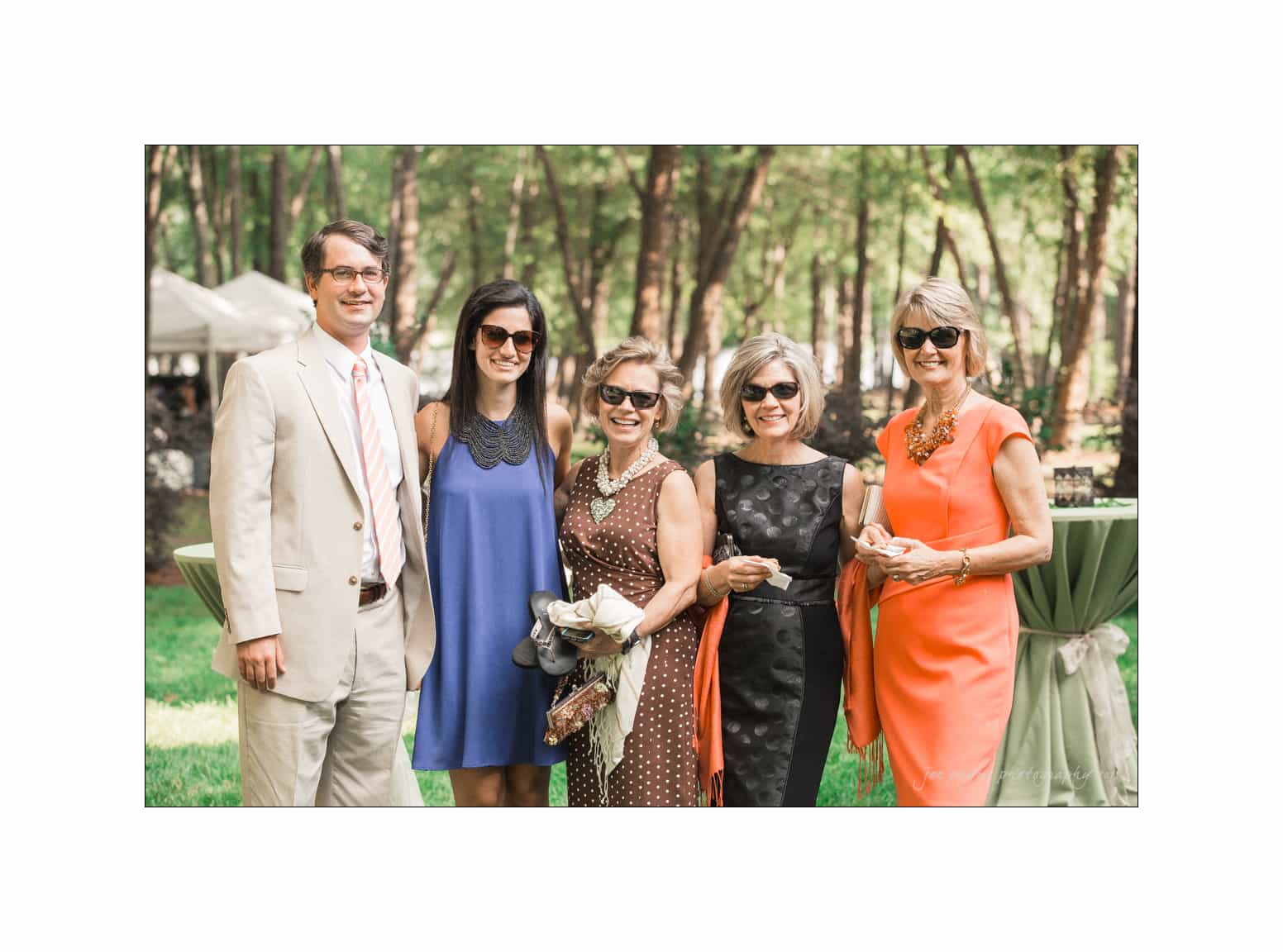 NC wedding photography - rachel & joe A group of five people dressed for a formal outdoor event stand together, smiling at the camera. They are wearing sunglasses, and the background is a lush, green, wooded area with sunlight filtering through the trees.