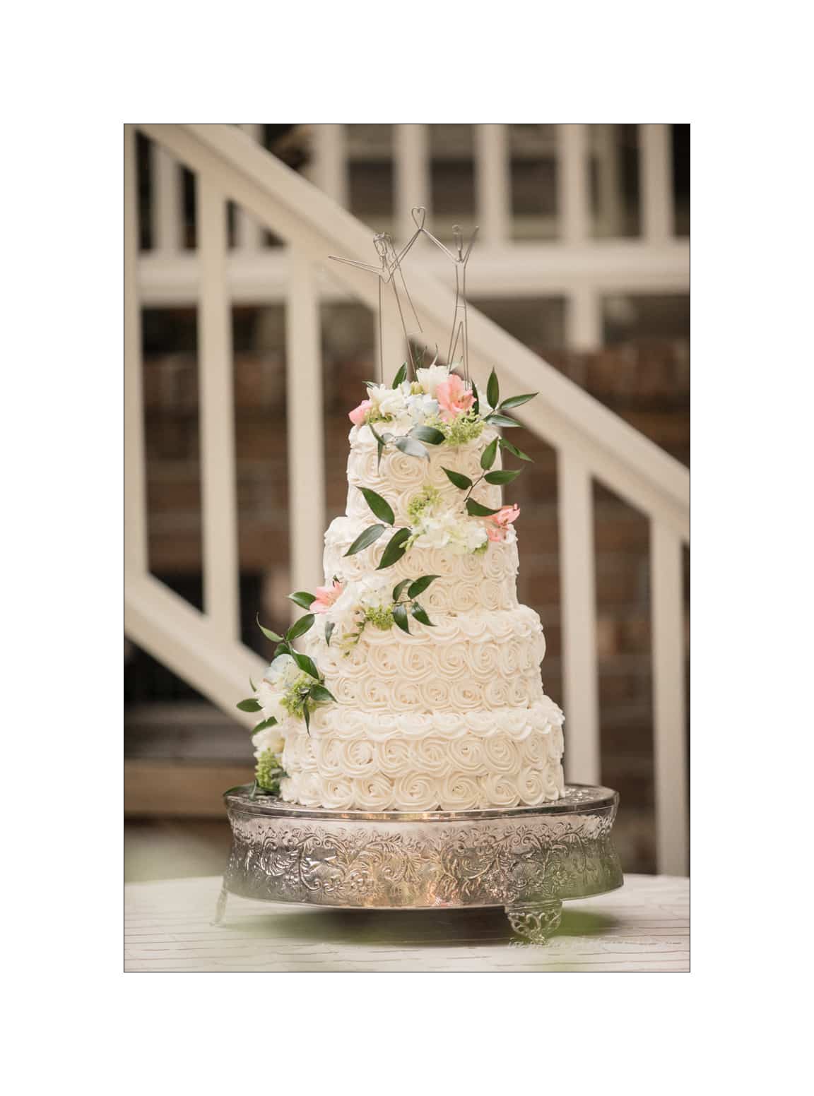 NC wedding photography - rachel & joe A three-tier white wedding cake adorned with pink and white flowers and greenery, displayed on an ornate silver stand. The background features a white staircase and wooden brick walls.