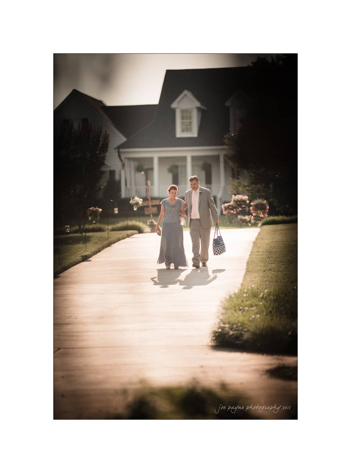 NC wedding photography - rachel & joe A couple walking hand in hand along a sunny path towards a large, elegant house in the background. The woman is wearing a long dress and the man is in a suit, carrying a patterned bag. The scene is warmly lit, suggesting late afternoon.