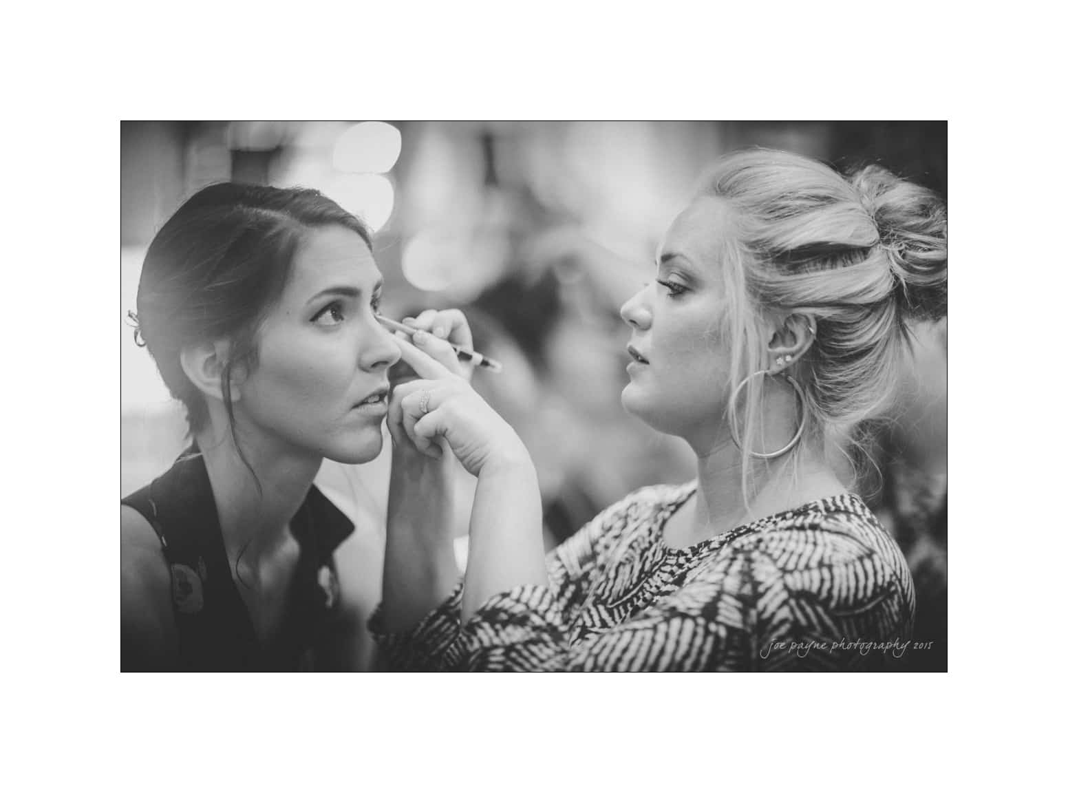 NC wedding photography - rachel & joe A woman with her hair in a bun applies makeup with a brush to another woman, who is looking ahead. The scene is captured in black and white, creating a classic and elegant atmosphere.