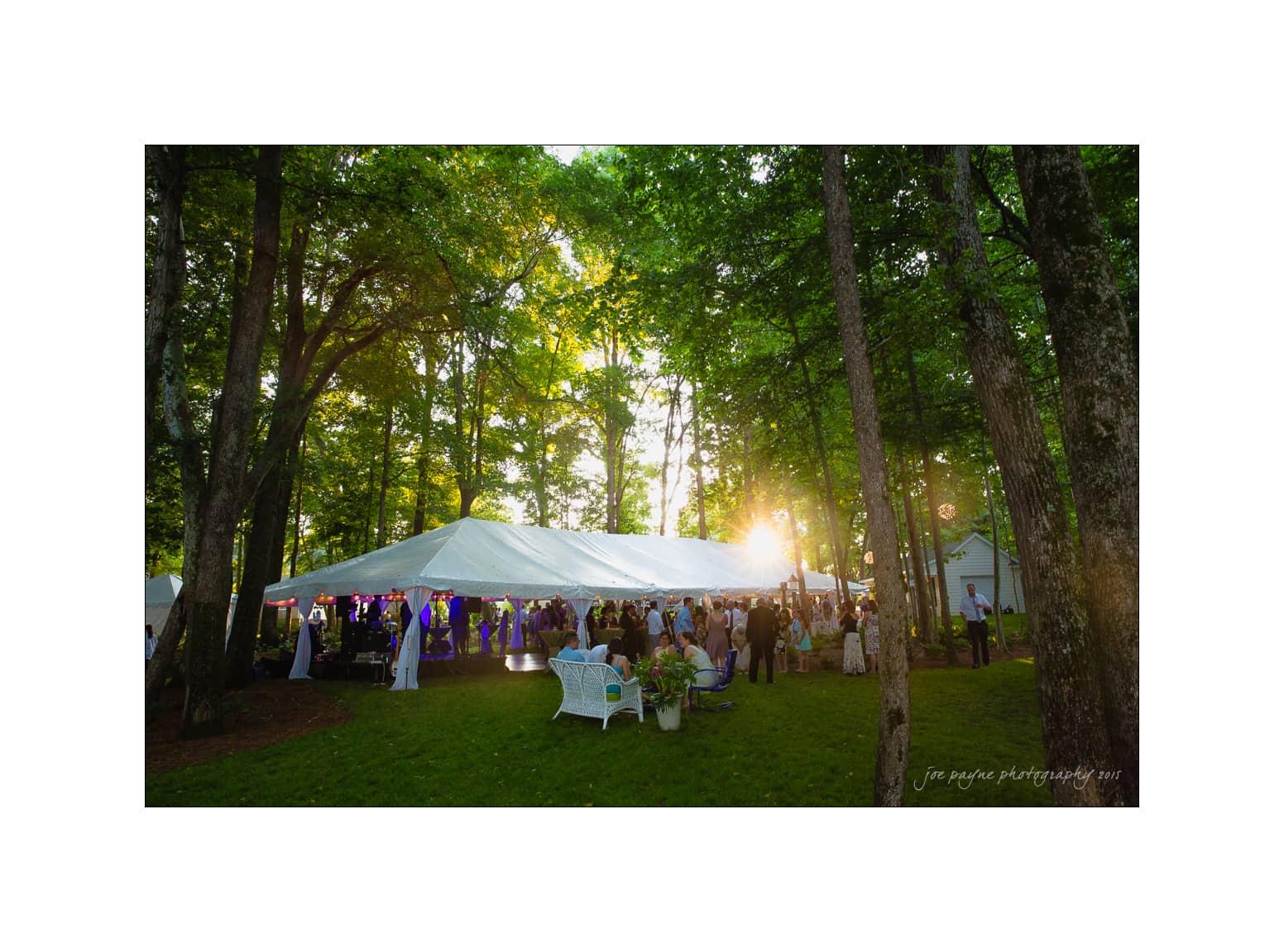 NC wedding photography - rachel & joe A wedding reception with guests mingling outside under a large white tent in a forested area. Sunlight filters through the trees, creating a warm, inviting atmosphere. A white bench is visible in the foreground.