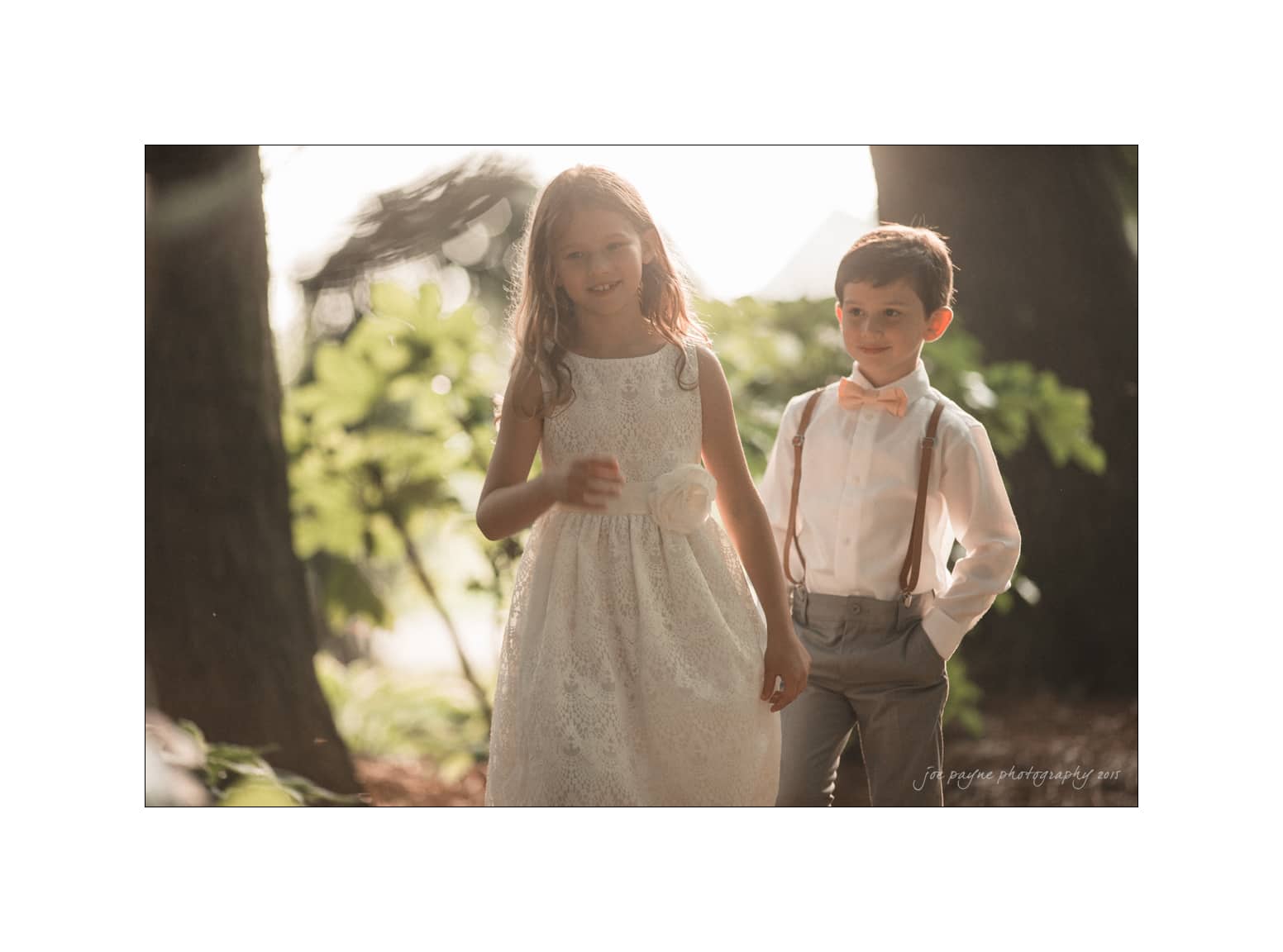 NC wedding photography - rachel & joe A young girl in a white dress and a boy in a white shirt with suspenders and a bow tie stand outdoors. The sunlight filters through the trees, creating a warm, soft glow around them.