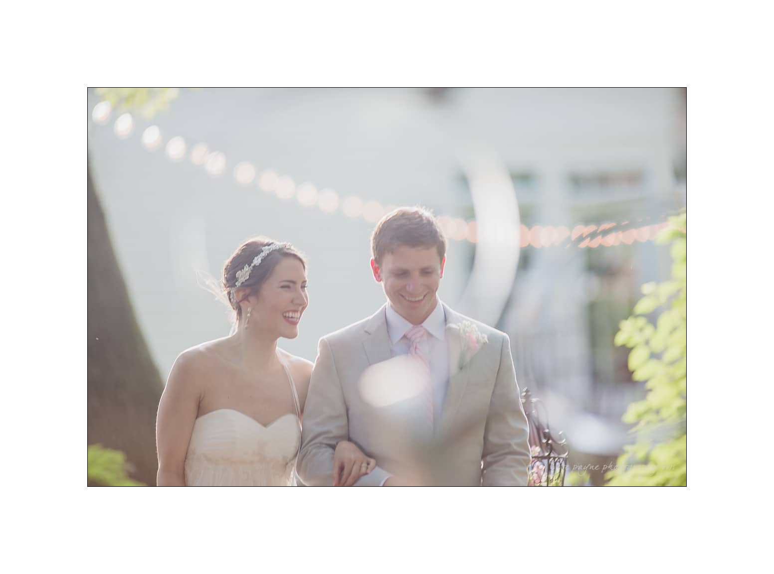 NC wedding photography - rachel & joe A couple dressed in wedding attire walks arm-in-arm outdoors. The bride wears a strapless white gown and a headband, while the groom wears a light suit with a pink tie. Soft focus and string lights create a dreamy atmosphere.