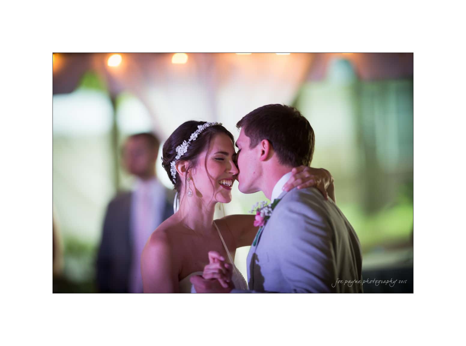 NC wedding photography - rachel & joe A bride and groom share a joyful dance at their wedding. The bride wears a strapless dress and a headpiece, and the groom is in a suit with a boutonniere. They smile as they embrace, with blurred lights in the background.