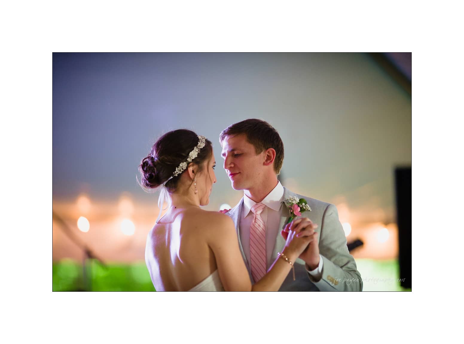 NC wedding photography - rachel & joe A couple in formal attire shares a dance, with the groom in a gray suit and pink tie, and the bride in a white strapless gown with a floral hair accessory. The background is softly lit, creating a romantic ambiance.