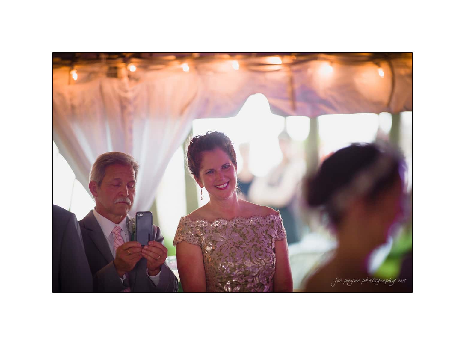 NC wedding photography - rachel & joe A woman dressed in an elegant dress smiles, while a man beside her captures a photo on his phone. They are indoors, with soft lighting and draped fabric in the background. Other people are partially visible, adding a celebratory atmosphere.