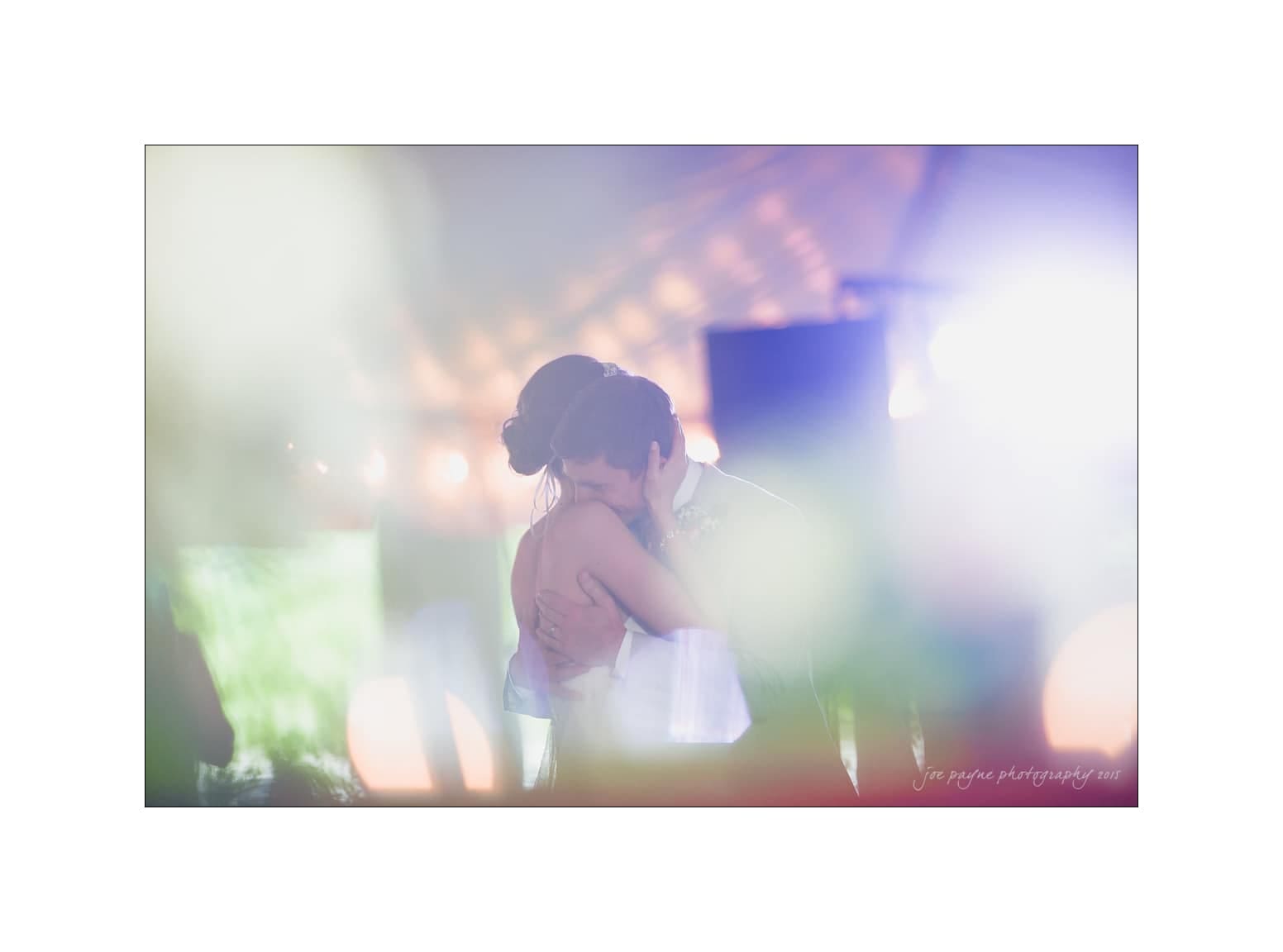 NC wedding photography - rachel & joe A couple shares a romantic embrace on the dance floor, enveloped in soft, colorful lighting. The background features blurred lights and a dreamy atmosphere, creating an ethereal scene.