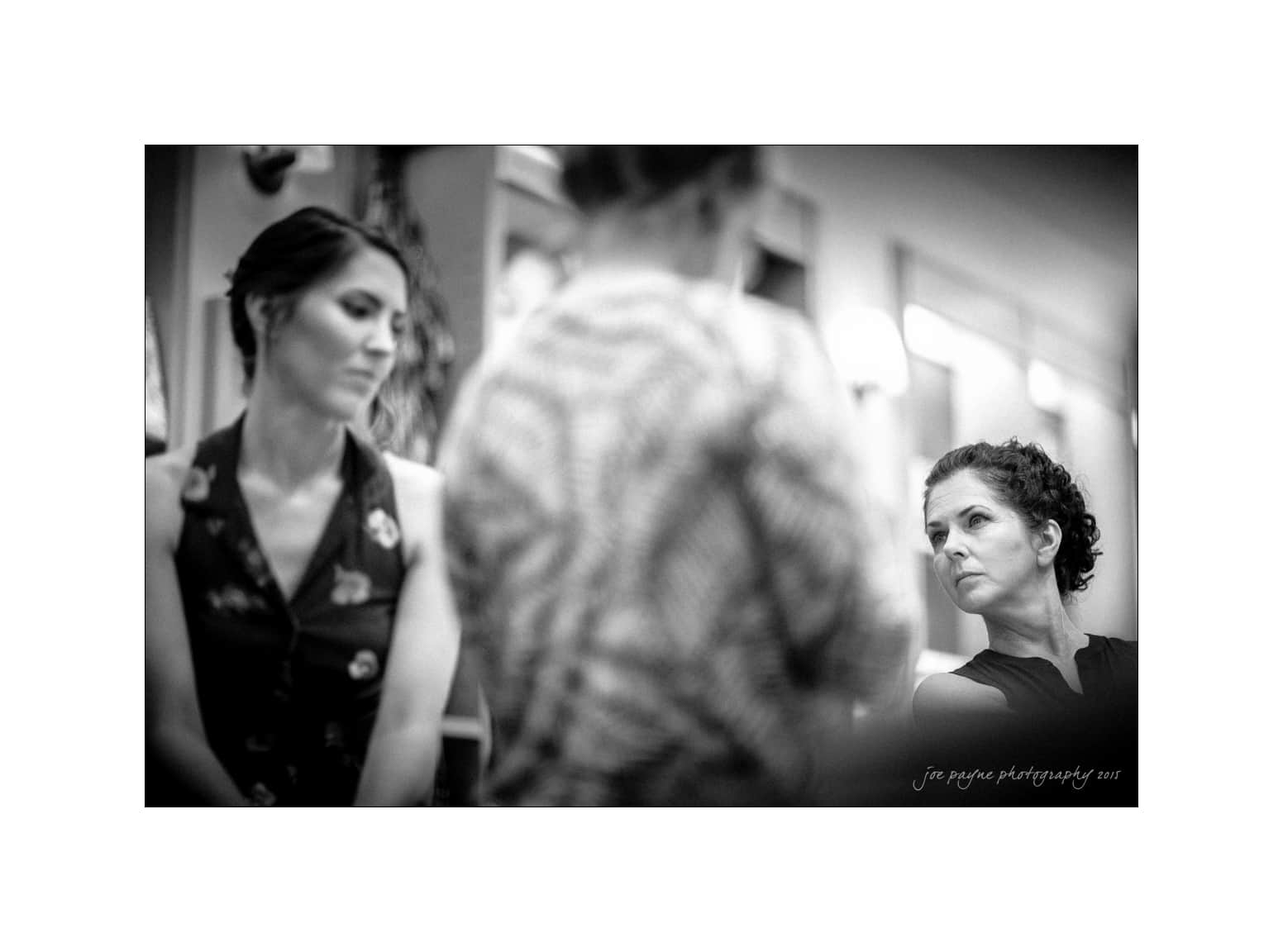 NC wedding photography - rachel & joe Black and white photo of two women in a room. One woman is seated, wearing a dark sleeveless top and looking to the side. The other woman, standing, is blurred in the foreground. The focus is on the seated womans expression.
